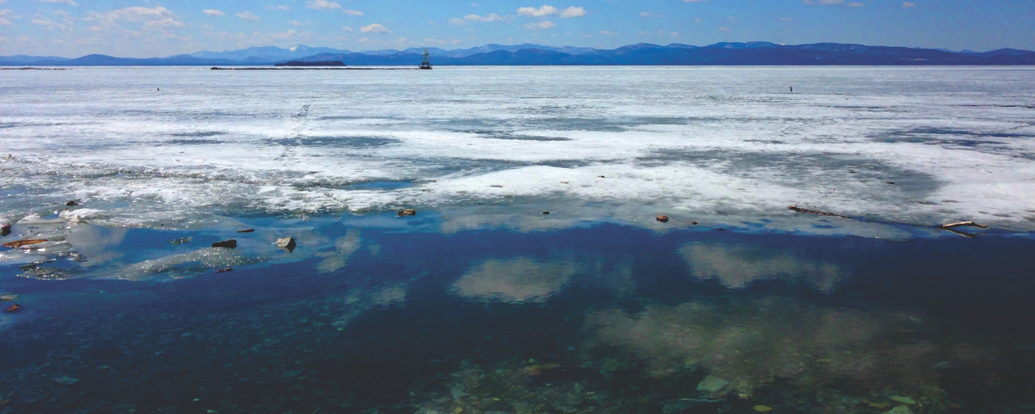A stunning photo of Lake Champlain during winter with blue mountains and a blue sky in the background and frozen icy water in the foreground.