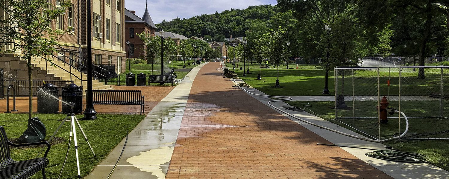 A photo of the completed Ellis Way pathway at UMass Amherst. The sun is shining, there are sprinklers going, and there's lots of green.