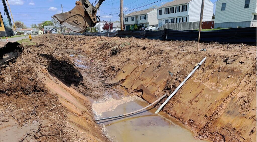 This image shows a waterline exposed in a ditch with brown water flowing underneath and a large bucket truck hovering over it. Finding buried hidden utilities was part of this safety improvement project.