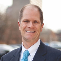 Headshot of Matthew Low standing outside in a blue blazer and light blue tie. Buildings in the background.