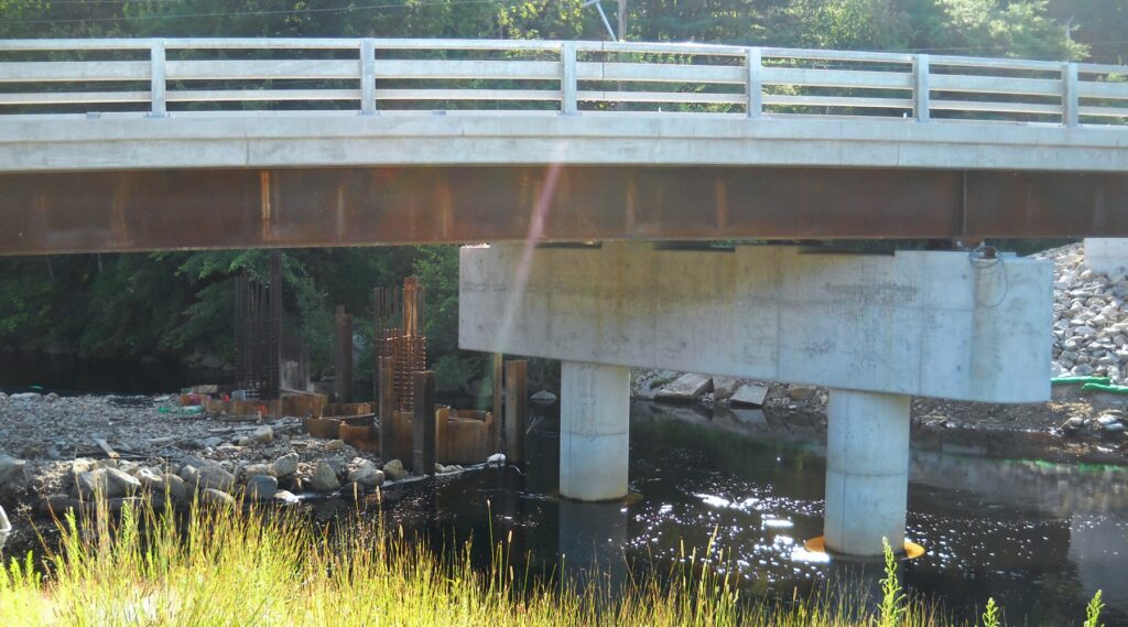 A photo focusing on the piers of the bridge construction. Photo showcases water, some green grasses in the foreground, and a lens flare.