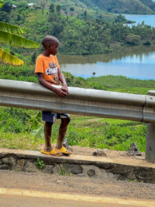 A photo of a young boy in Rwanda holding onto a guardrail with beautiful Rwandan greenery and water in the background. 