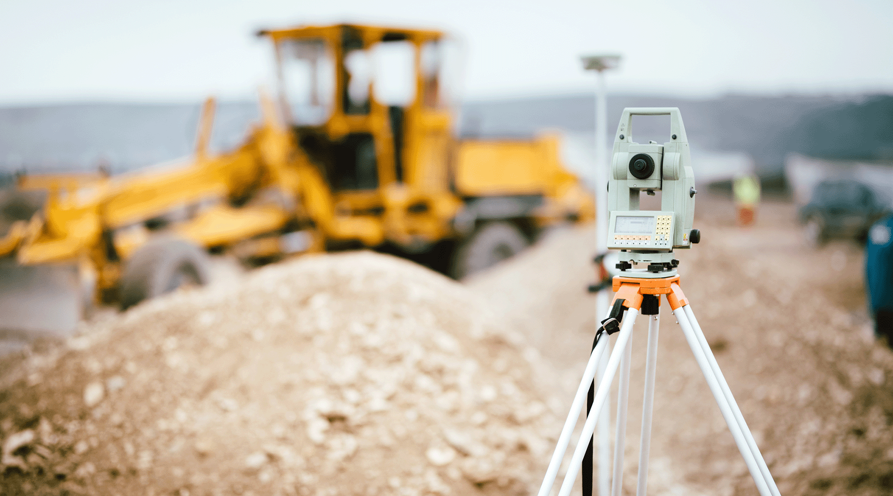 Featured image is a survey tool pointed towards a yellow cat machine in the background of a construction site.