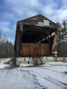 An image of the Whittier Covered Bridge from the front, removed from its location over the river to be rehabilitated. There is snow on the ground around the bridge.