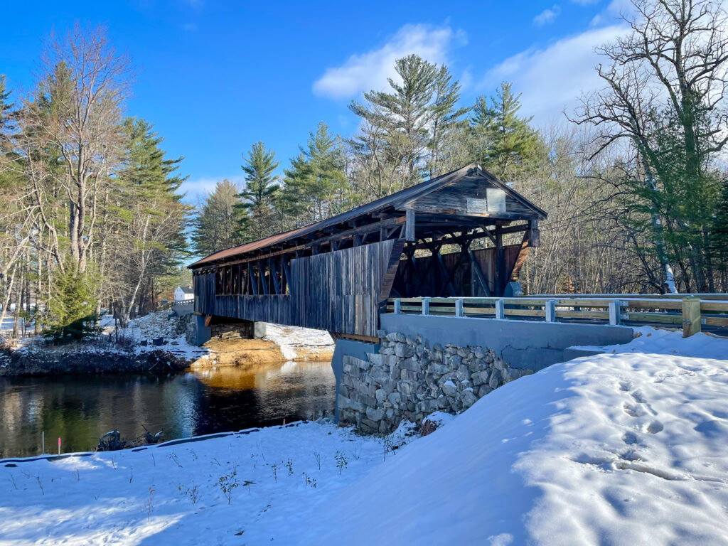 A profile view of the Whittier Covered Bridge back over the river with its new abutments. There is still snow on the ground and there is a bright blue sky.