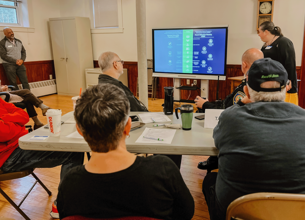 An image of someone presenting with Dunstable citizens sitting in chairs facing the presentation. The photo shows the back of peoples' heads and the screen.