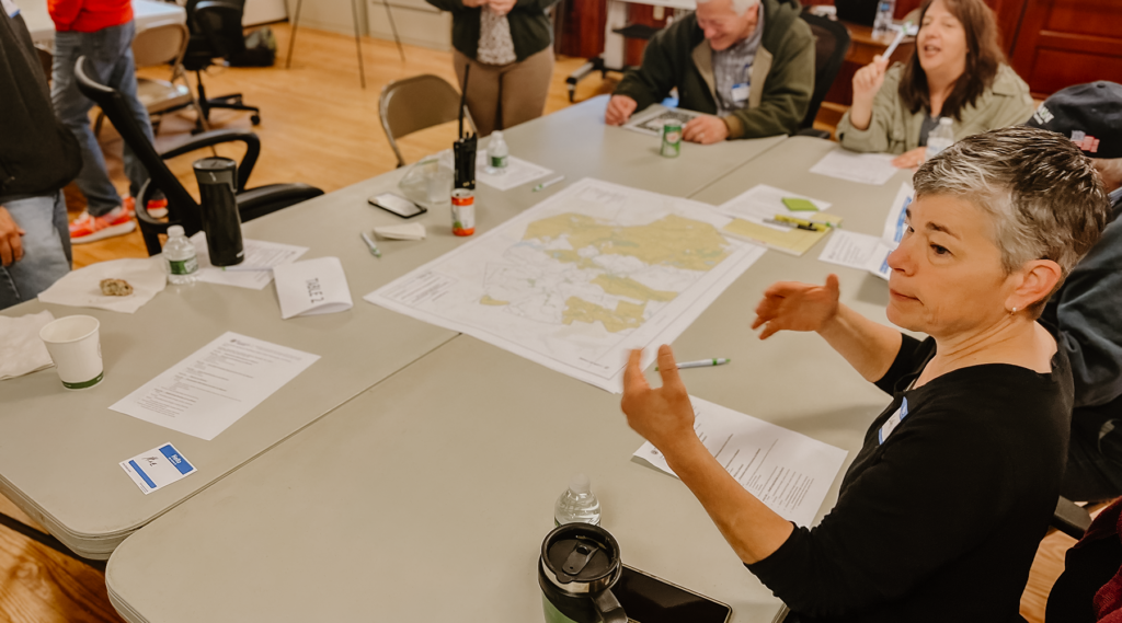 A woman is sitting at a table with plan sets spread out. Other people sit around the table but you can't see their faces. The woman in the forefront of the photo is speaking and her hands are raised.