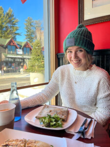 Mia Dischiavo - a photo in portrait orientation with Mia sitting down at a restaurant with a plate of food in front of her. She's wearing a white sweater and a green beanie hat.