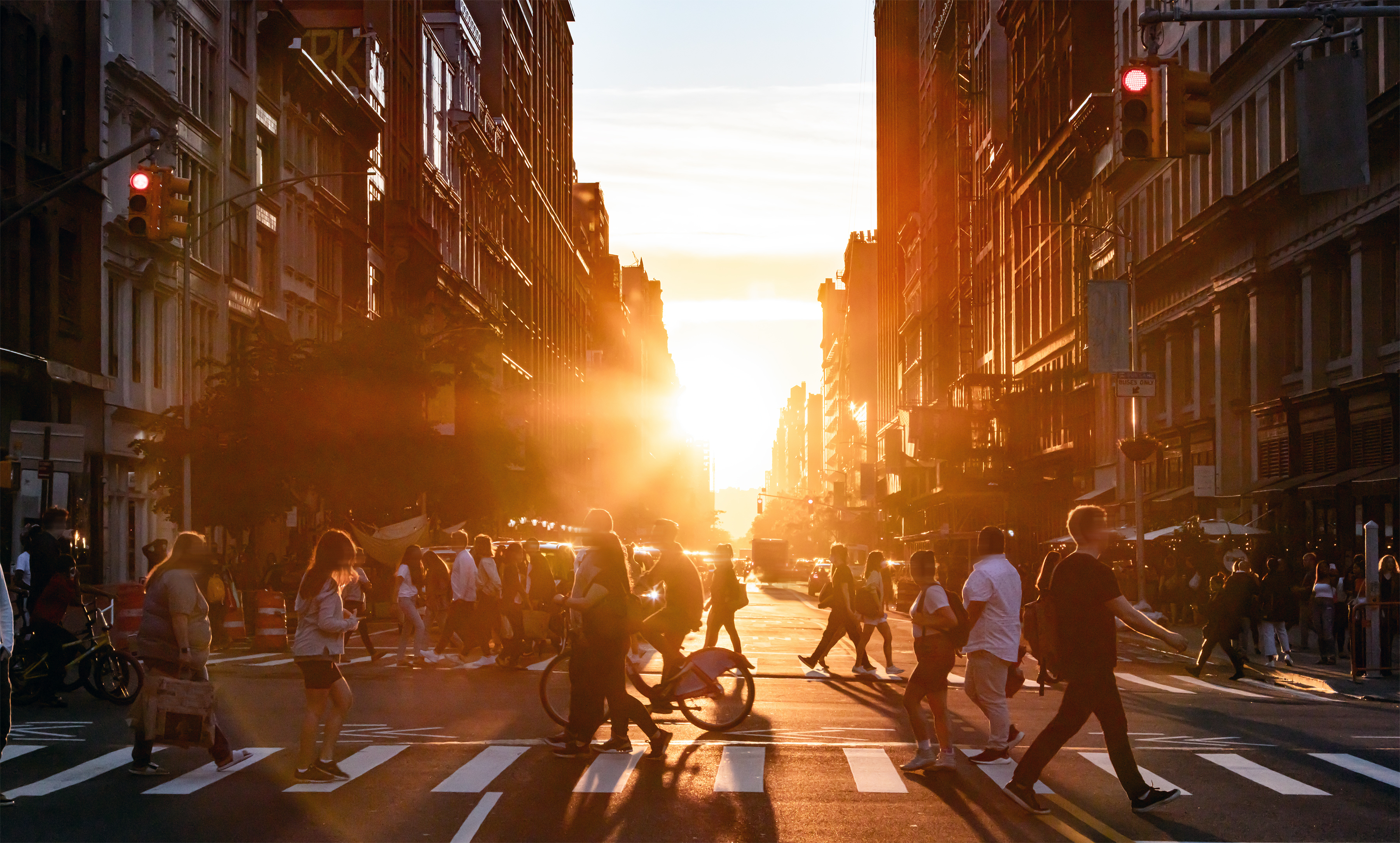 People walking through the busy intersection at 5th Avenue and 23rd Street in New York City on a summer day with sunset flare behind the background buildings