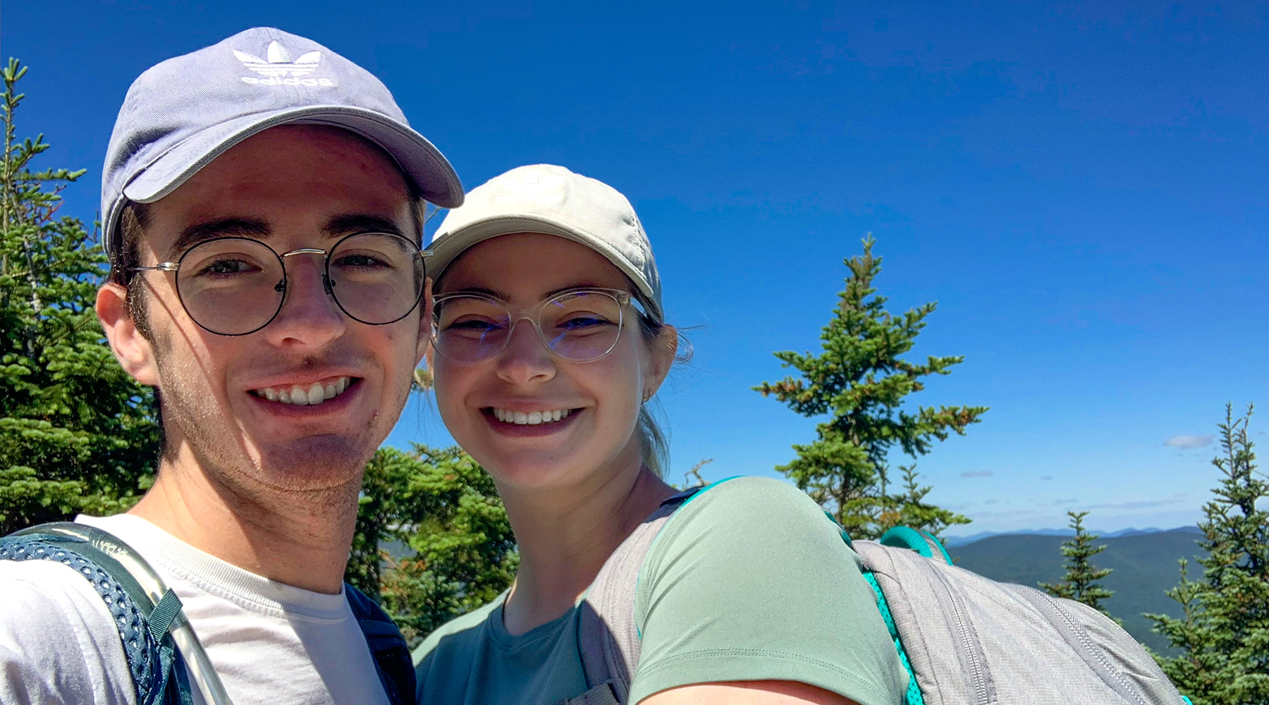 Allyson Kitchens is featured in a photo with a man standing on top of a mountain. They are smiling at the camera. Bright blue skies are behind them.