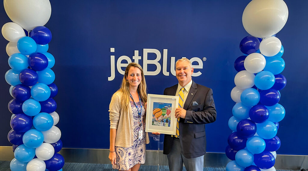 An image of Bob Furey on the right holding a sign that says Presque Isle. To his left is a woman from the Presque Isle Airport. They are both standing in front of a blue wall that says JetBlue.