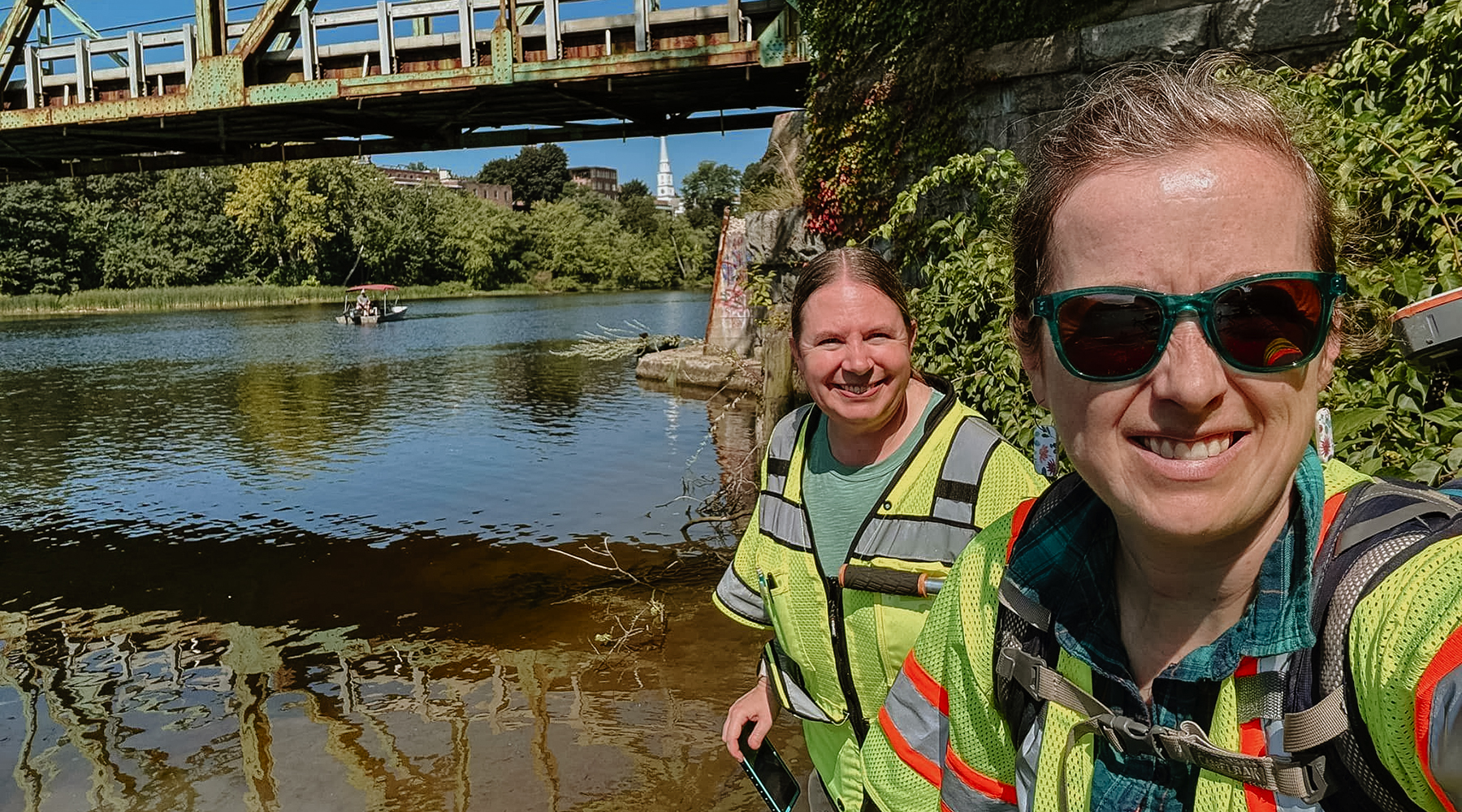 A photo of Melinda and Joanne taken while near a bridge outside. They are smiling into the sunshine at the camera. Both are wildlife biologists.