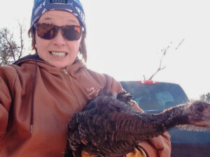 Melinda is in a brown sweatshirt and sunglasses. She's taking a selfie while holding a live turkey.