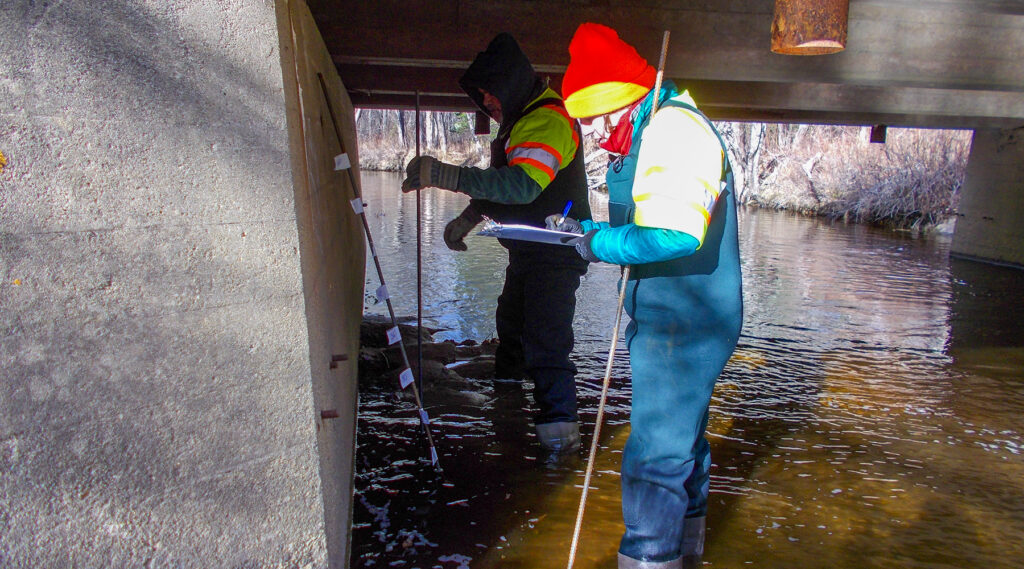 A photo of Lynne with waders on in water inspecting scour for a bridge.