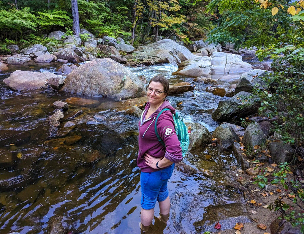 Karen Allen's employee spotlight photo. Karen has a bright blue backpack on and is standing with bare feet in a mountain stream.