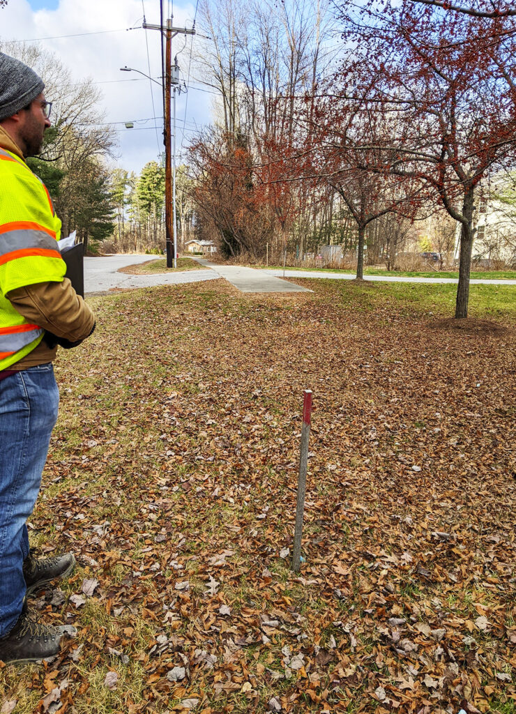 Jon Olin, wearing a bright yellow safety vest, is at the edge of the photo looking out into the roads. The ground is covered in fall leaves and there are red-leafed trees in the background.