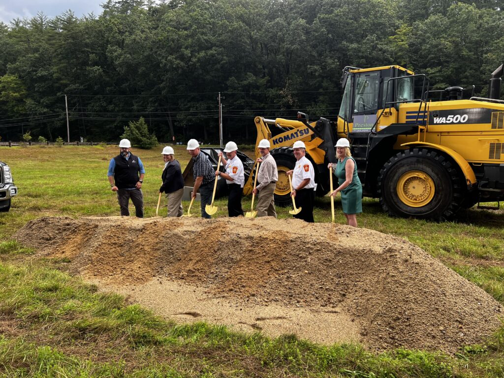 The ground breaking ceremony at New Boston Fire Station features officials and Kimberly Peace digging in a dirt mound with shovels and a large yellow machine behind them.