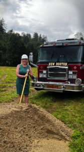 A photo of Kimberly Peace posing in a hard hat while holding the ground-breaking shovel. A New Boston fire truck is in the background and it's cloudy outside.