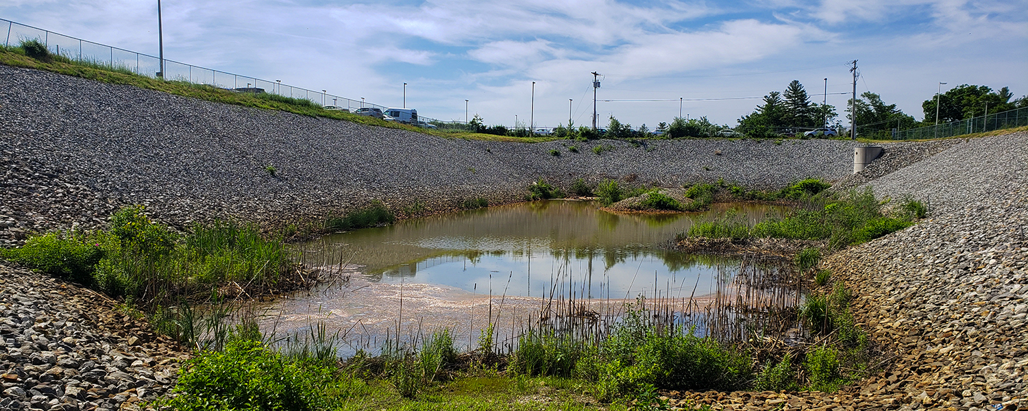 A drainage area at Manchester-Boston Regional Airport. The photo looks over a pond below road level. Blue skies surround the infrastructure.