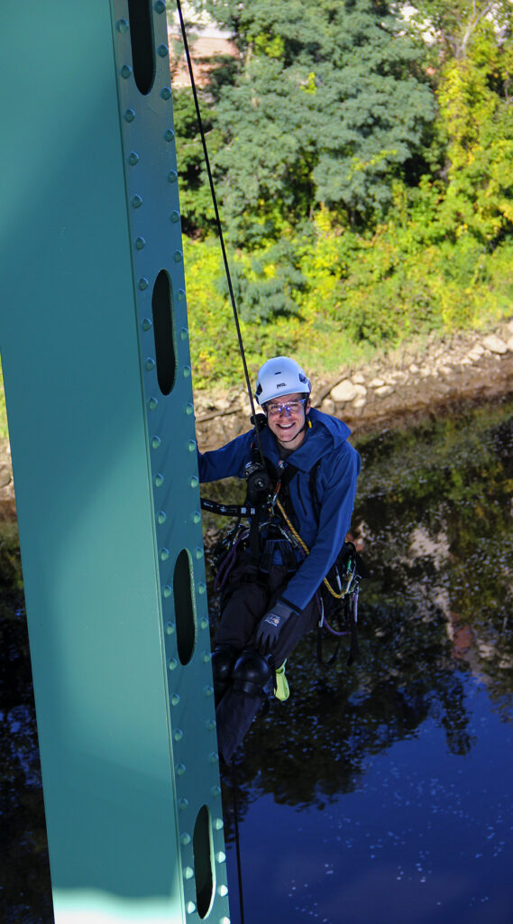 Joe Ripley doing bridge inspection over water. He is wearing ropes and smiling at the camera on a sunny day.