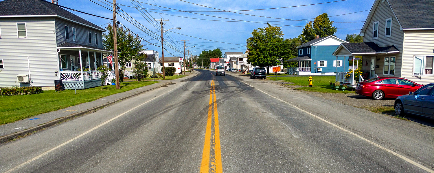 A photo taken in the middle of the road looking at the newly paved sections in Van Buren, ME. The sky is blue and the sun is shining. There's a clear divide between fresh and old pavement.