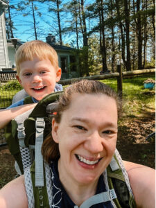 Megan Ooms spending time with her son, who is on a backpack on her back. It's sunny outside and they are smiling at the camera.