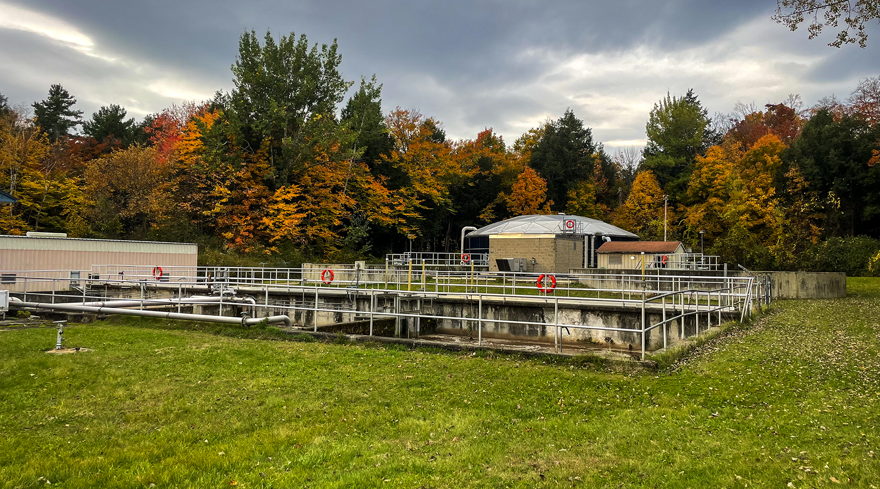 A photo of the Bartlett Bay wastewater treatment facility in the fall. Foliage is behind some of the equipment and the sky is cloudy.