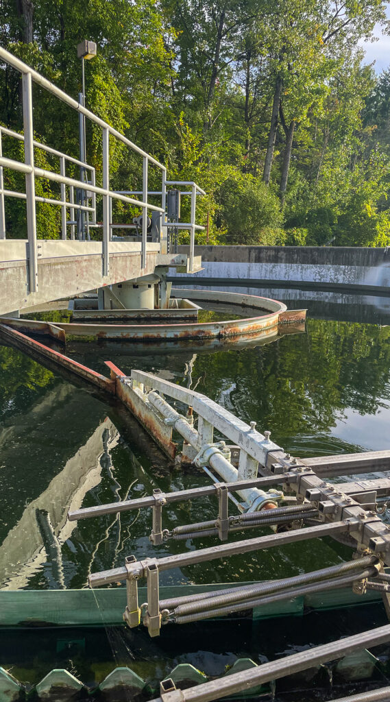 A photo of the clarifier at the Bartlett Bay wastewater treatment facility. The clarifier is outside and there is green foliage all around and reflecting in the water.