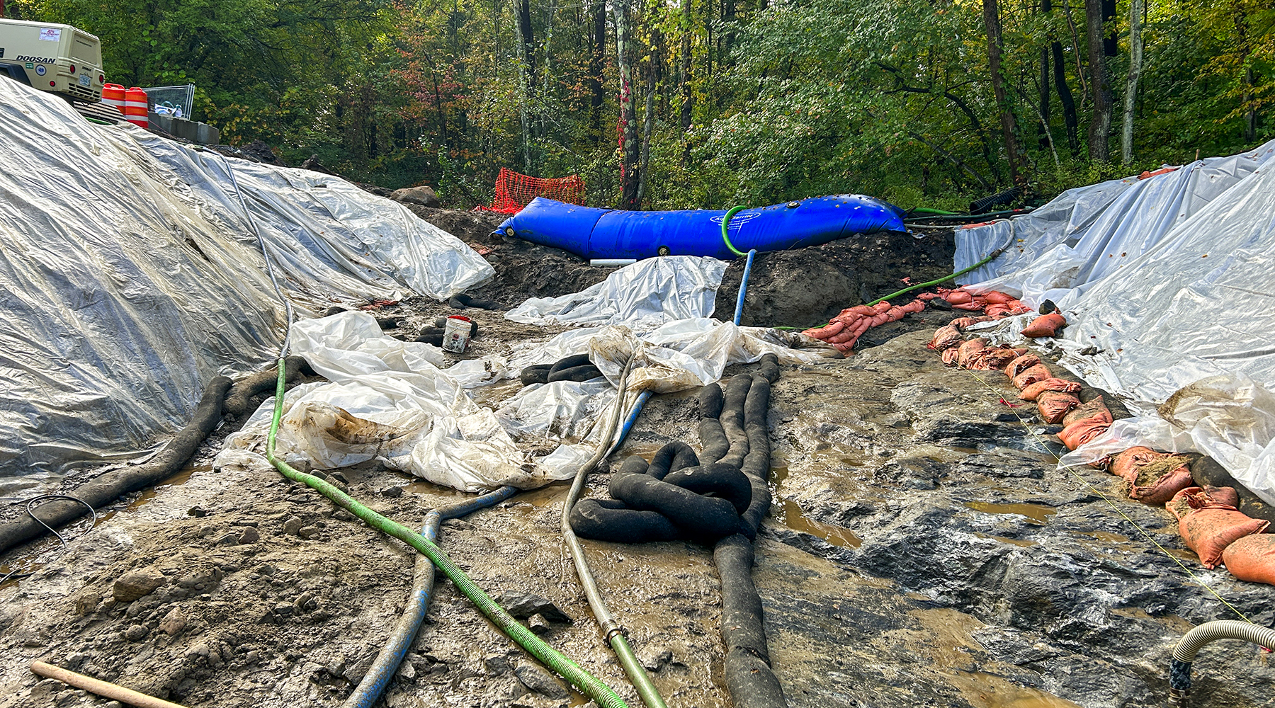 An image of the construction site. Tubes lay all over a muddy ground and plastic-covered slopes are on both sides of the photo.