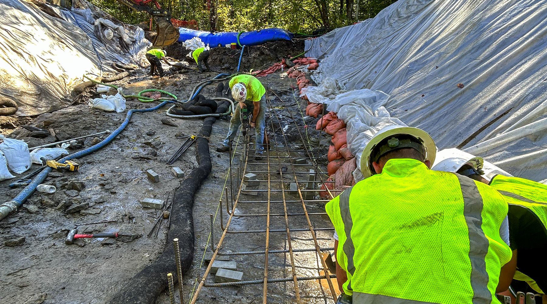 A photo at a construction site with construction crew kneeling down and working on the ground. Slopes are on both sides, covered in large sheets.