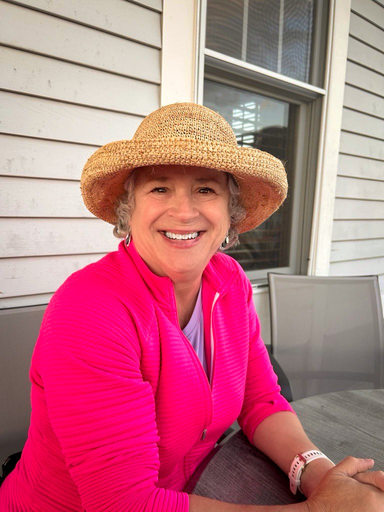 A photo of Angie in portrait mode. She's wearing a straw hat and a bright pink active sweatshirt. She's sitting down outside smiling at the camera.