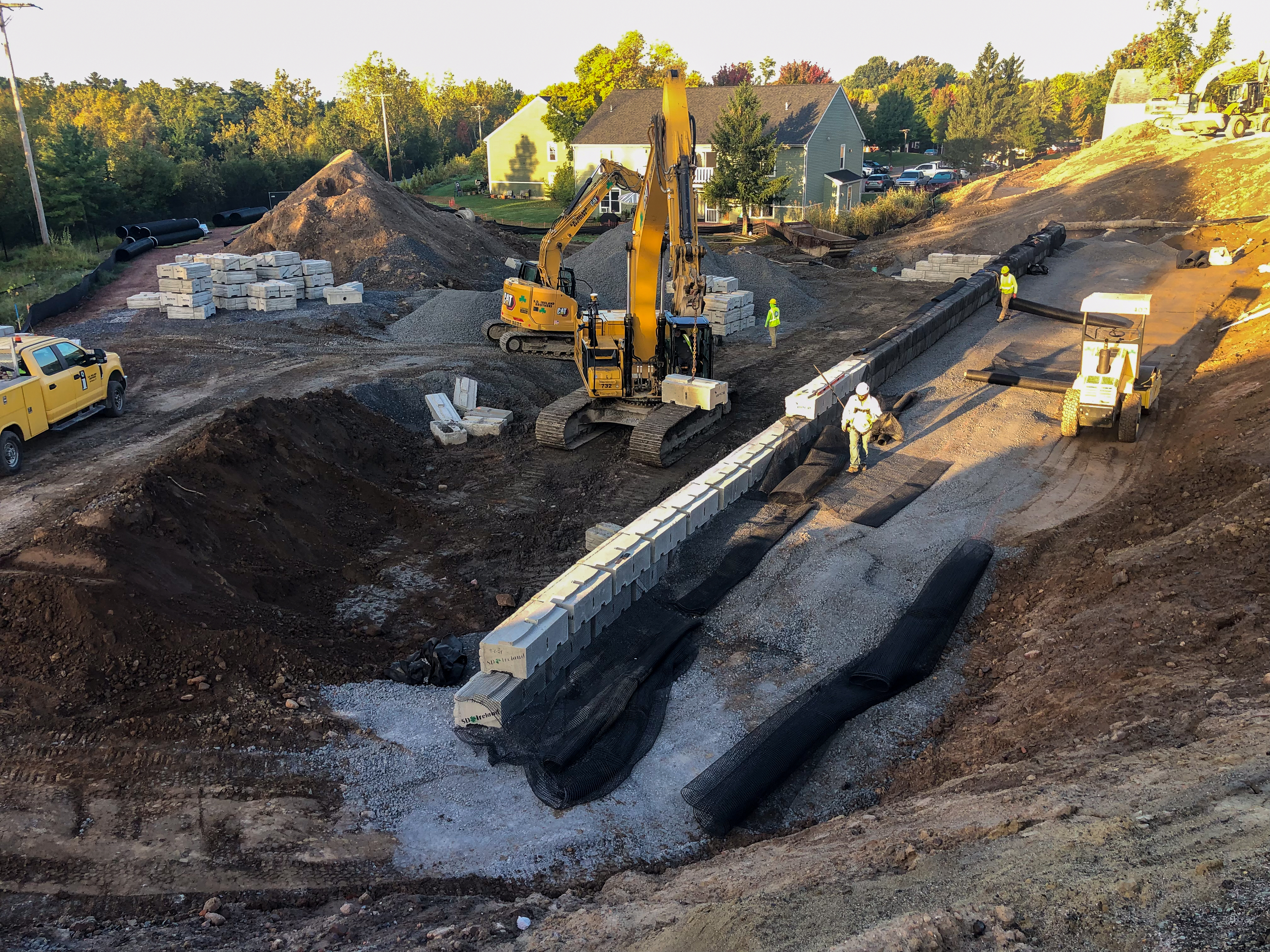 "Construction site with workers installing a retaining wall using large concrete blocks. An excavator and a compact roller are in use, while workers in safety gear handle geotextile fabric. Stacks of materials and construction vehicles are visible in the background near residential houses.