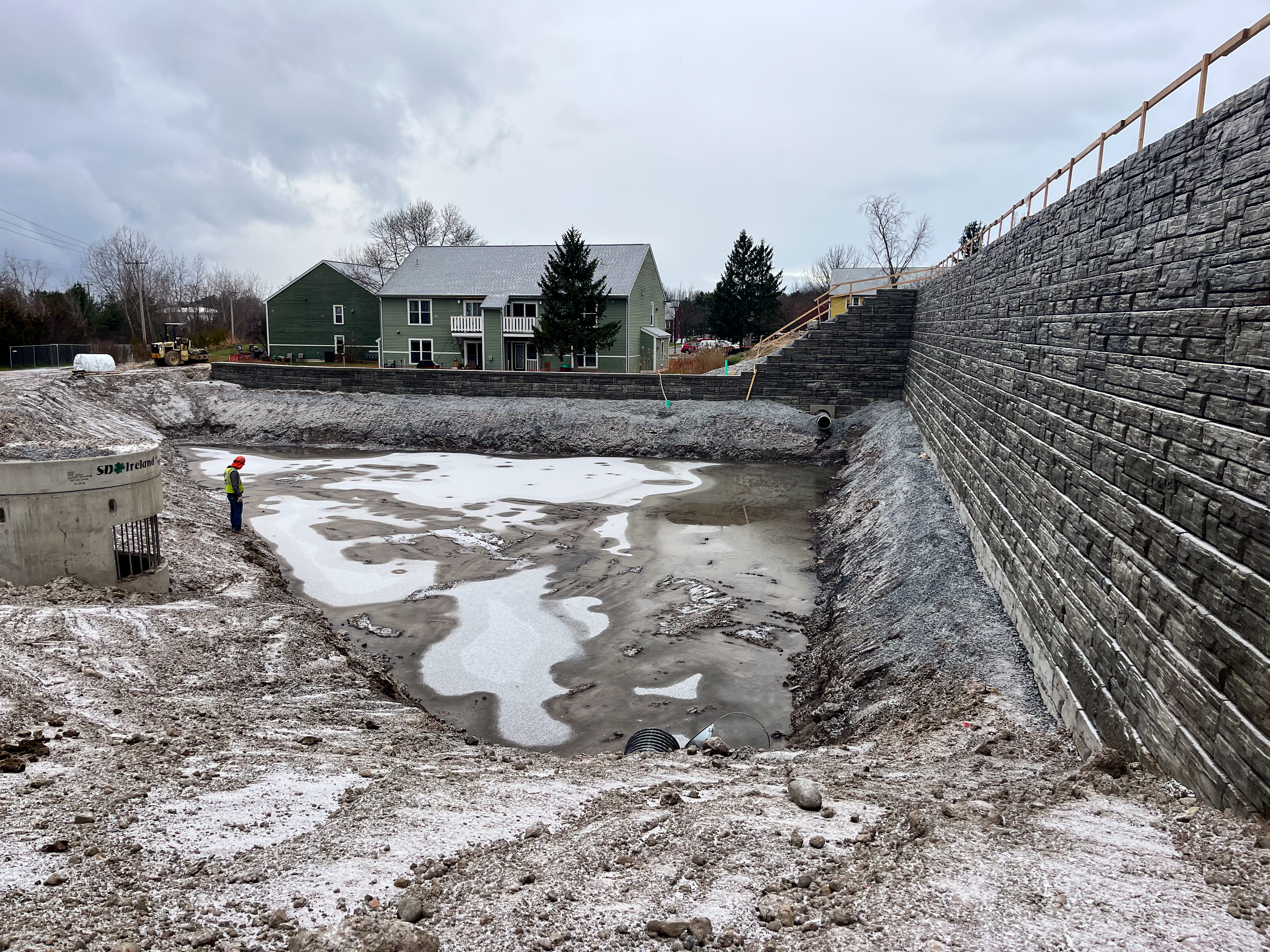 Construction site featuring a large retaining wall and a partially frozen stormwater retention pond. A worker in safety gear stands near a concrete drainage structure labeled 'SD Ireland.' Residential houses and construction equipment are visible in the background under an overcast sky.