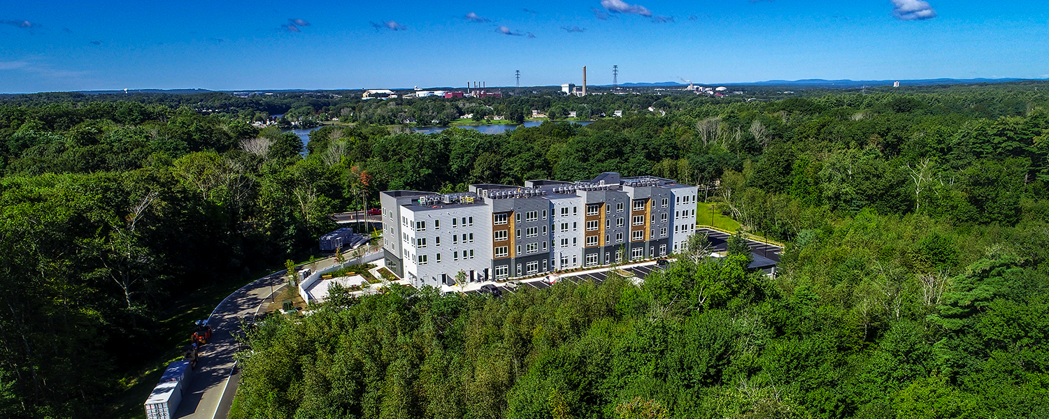 Aerial image of the Kittery project from a distance. You can see green forest all around the property and bright blue skies on the horizon. The building is new.