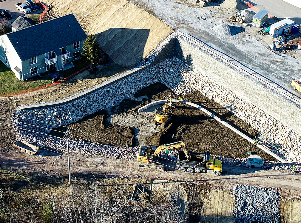 Aerial view of a construction site featuring a large retaining wall made of stacked stone and concrete blocks. Excavators are working on grading and placing materials, while a dump truck and workers in safety gear are present. A nearby house is protected by a silt fence, and construction equipment is visible in the background.