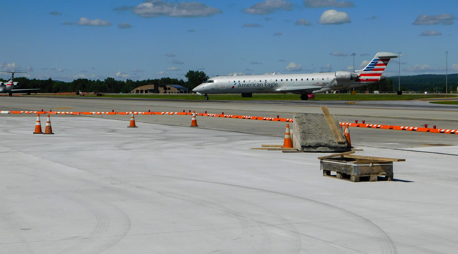 A photo featuring airport pavement and an airplane in the background. The sky is mostly sunny and blue with a few clouds.