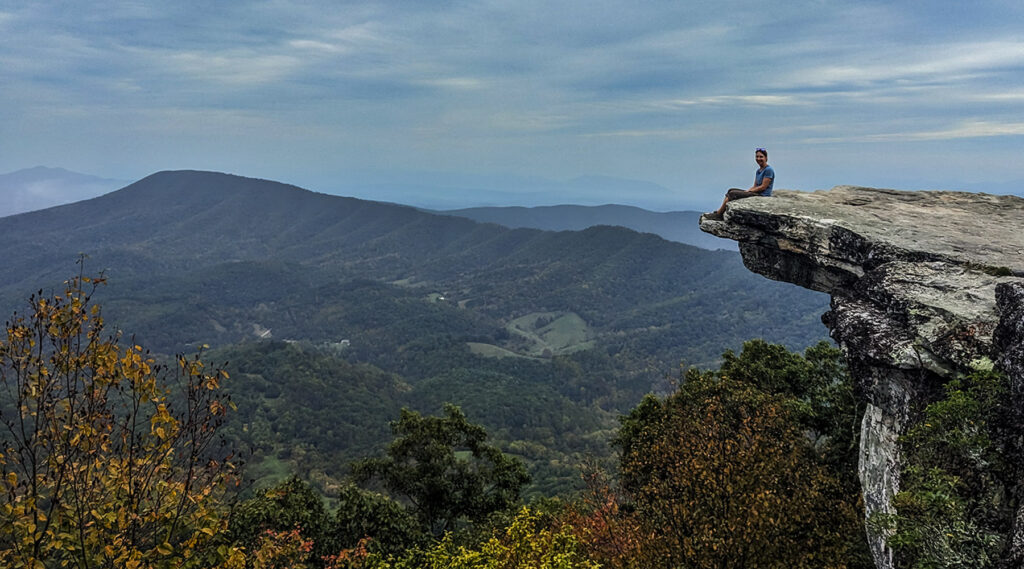 A photo of Jenae sitting on the edge of a cliff surrounded by mountains and fall foliage.