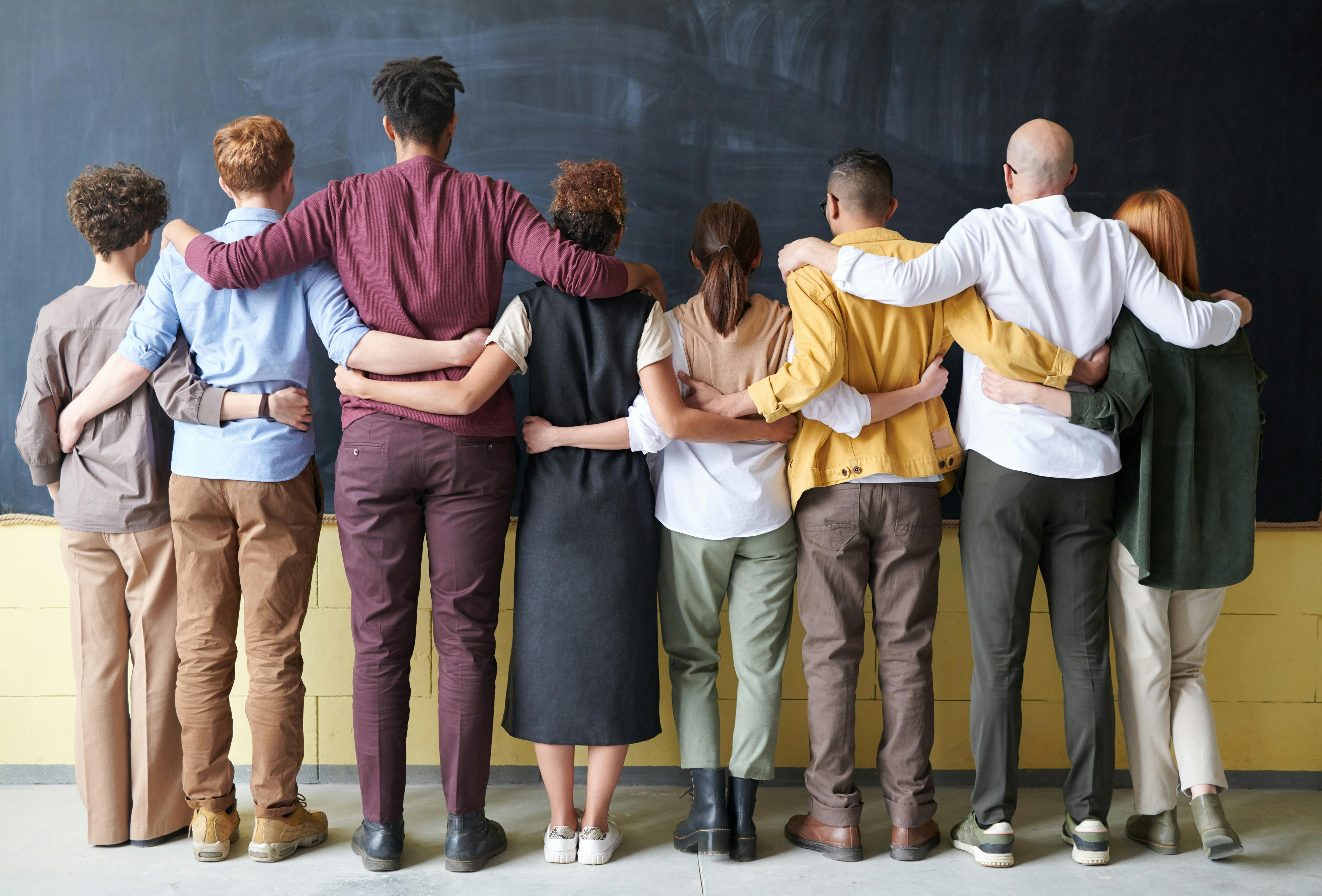 A diverse group of eight people standing in front of a chalkboard with their arms around each other, viewed from behind, symbolizing unity and support.