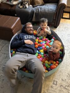 Eric Doe is wearing a UNH sweatshirt and is playing with his children inside his home.