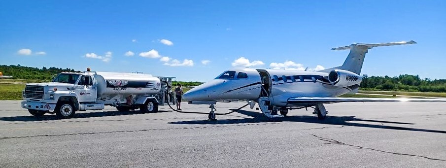 A private jet at Millinocket Airport. The jet is parked and is getting refueled by a truck. It's a sunny day.