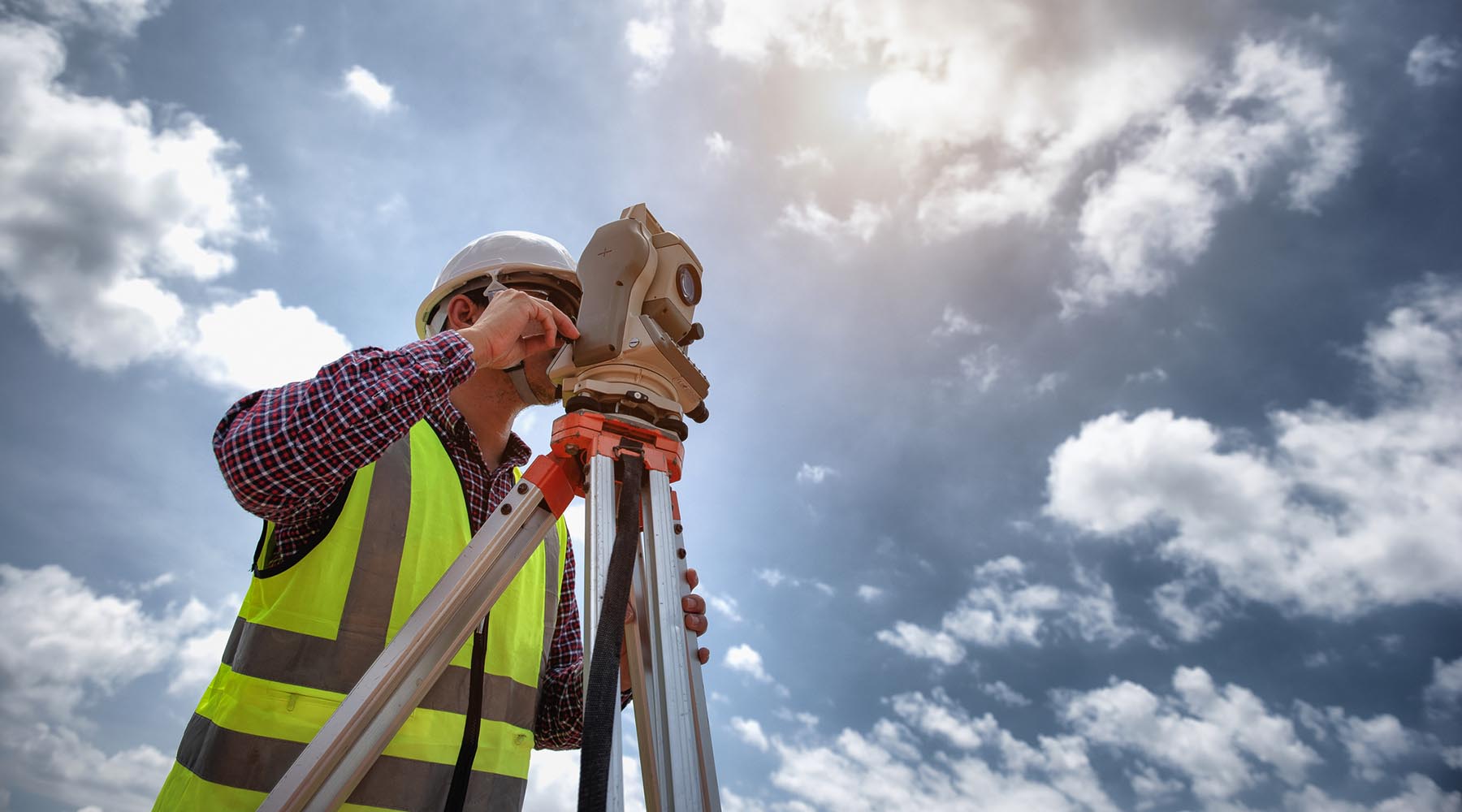 Survey image with a surveyor with their equipment and bright blue sky and clouds behind them