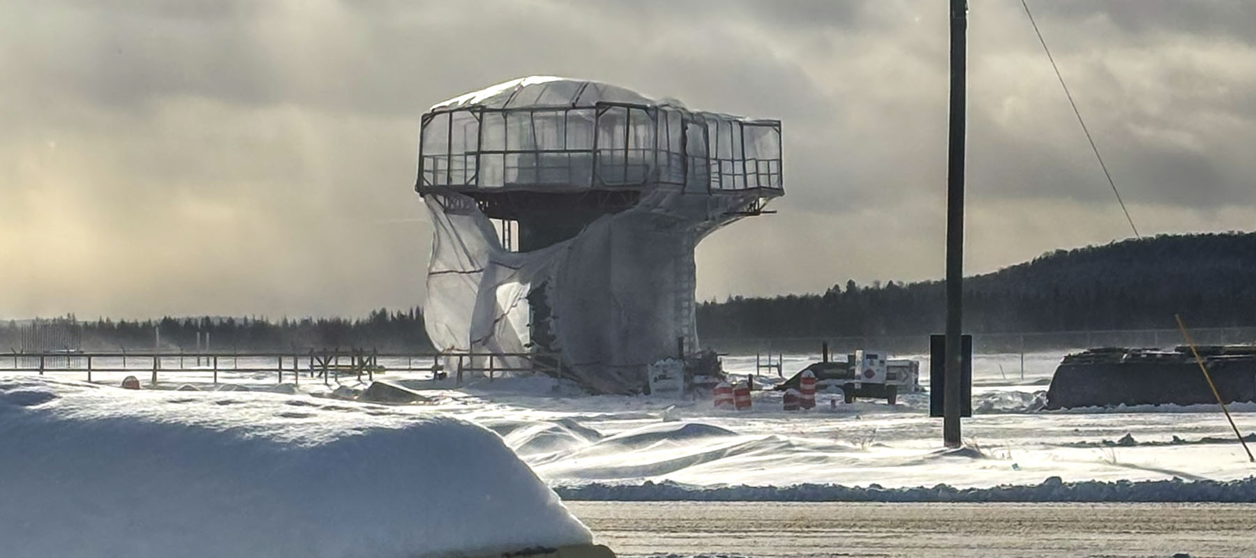 Another photo of airport infrastructure in winter that is somewhat constructed. There is plastic sheeting on a small tower and snow all around. It also looks hazy and blustery.