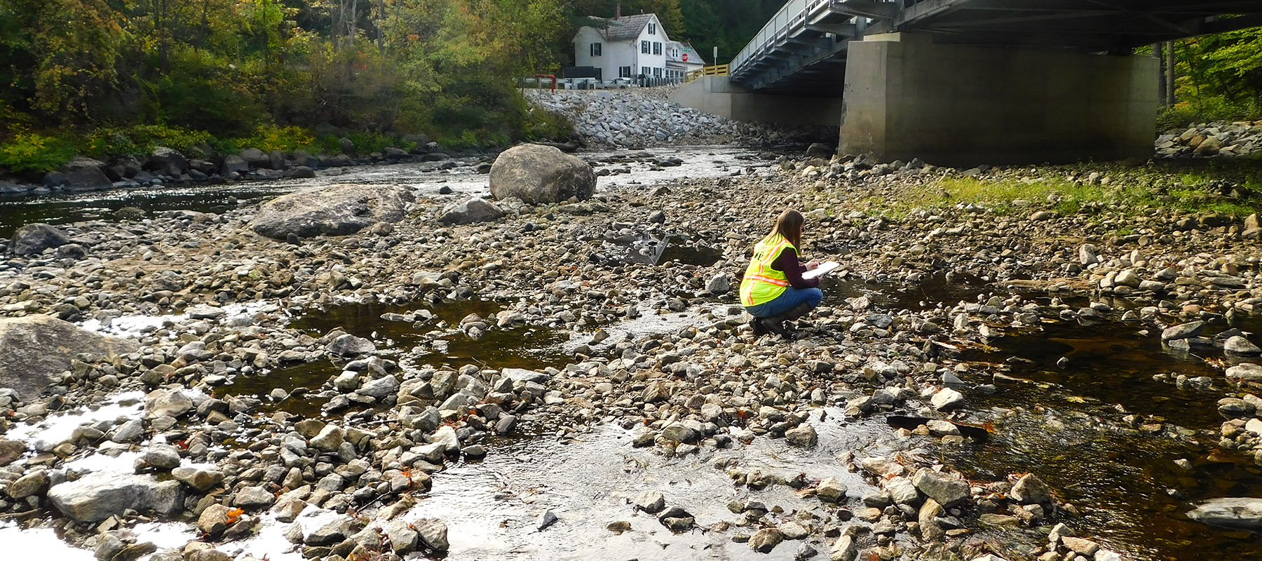 Joanne Theriault, an experienced environmental permitter, is out in the field wearing a safety vest, crouching in a stream near a bridge.