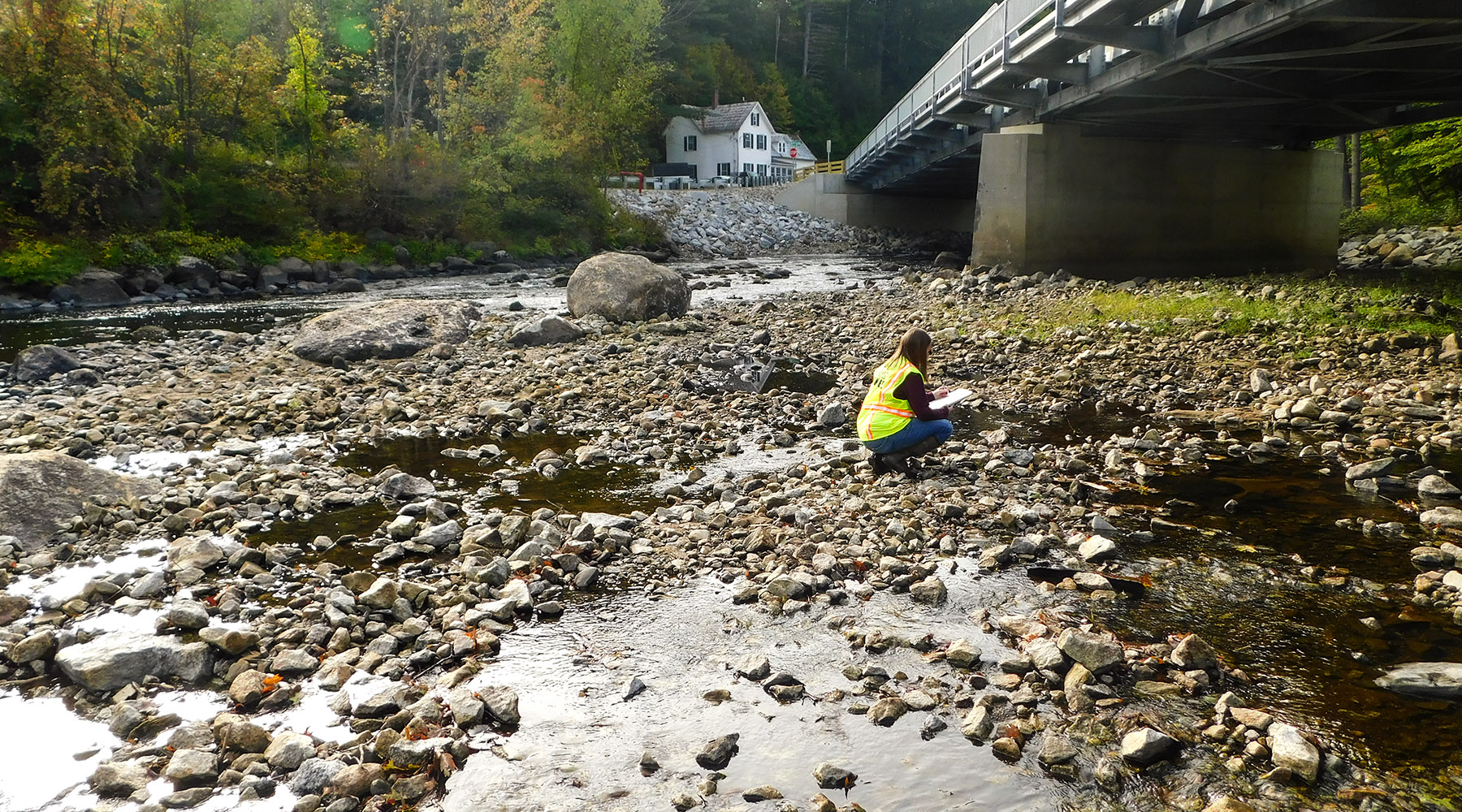 Joanne featured doing environmental permitting. She's wearing a high-vis vest in a stream with a bridge nearby.