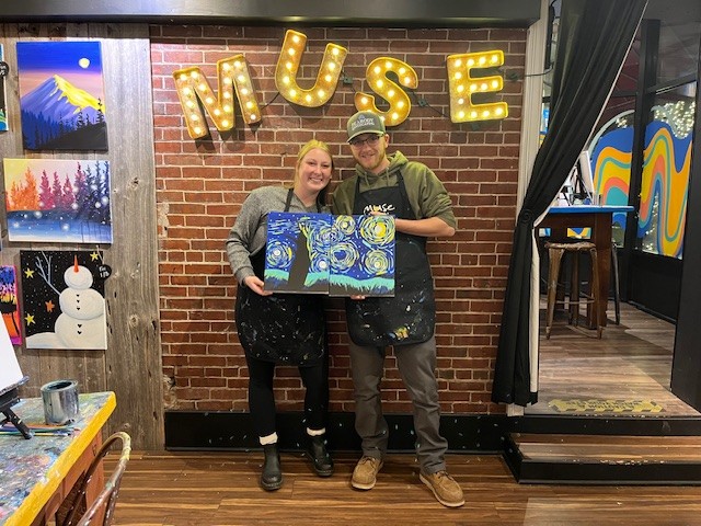 Bailey Jackson featured with her partner during a paint night at Muse Paintbar. They stand smiling in front of the Muse sign holding their paintings.